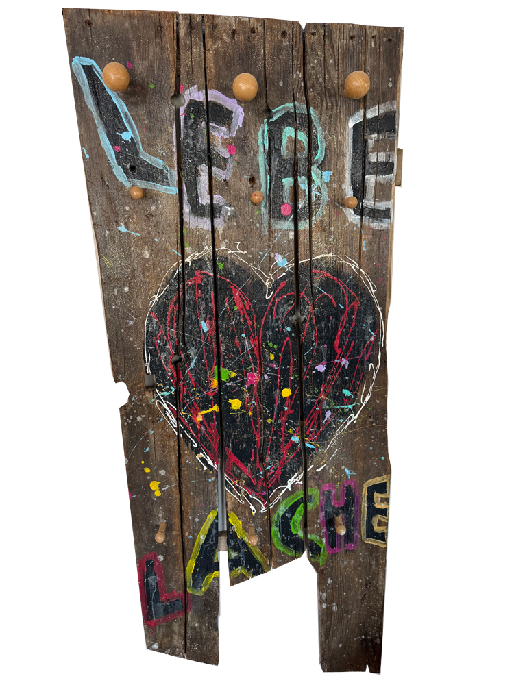 Skulpturale Urban-Pop-Art-Garderobe aus alten, verwitterten Holzbrettern. Im oberen Bereich steht das handgemalte Wort „LEBE“ über runden Kleiderhaken aus Holz, im Zentrum befindet sich ein großes, mit bunten Farbspritzern gefülltes Kreideherz. Am unteren, unebenen Rand ist das Wort „LACHE“ in bunten Buchstaben aufgetragen.